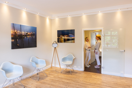 Zwei Personen stehen und unterhalten sich in einem gut beleuchteten Wartebereich. An der Wand hängen Stadtlandschaftsfotografien. Moderne Stühle und ein stilvolles Stativlicht ergänzen den Raum.