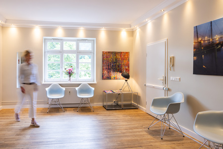 Eine Person geht durch einen hellen Warteraum. Kontext: Zwei Stühle vor einem Fenster, moderne Kunstwerke an den Wänden, Holzboden und weiches Licht schaffen eine einladende Atmosphäre.