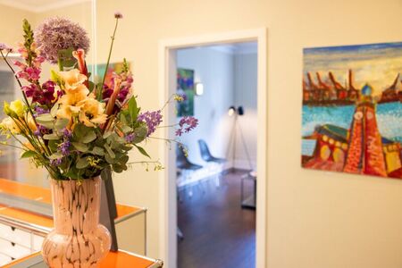 Ein bunter Blumenstrauß steht in einer Vase auf einem Tisch. Daneben führt eine offene Tür in einen Raum mit einem Stuhl und einem Stehlampen. An der Wand hängt ein farbenfrohes Gemälde.